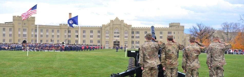 cadet battery saluting flags in front of barracks with corps in parade formation in background