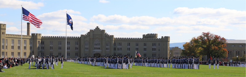 Cadets in parade formation on  fall day in front of barracks with flags blowing in the wind.