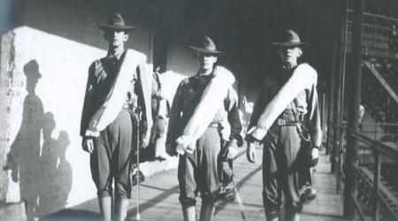Three cadets standing within barracks wearing a long white sash, pants tucked into their socks, and wide brimmed hats