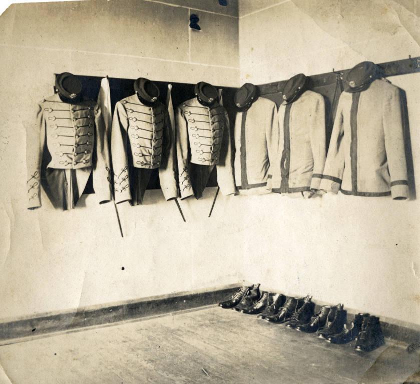 Three coatees and three grey blouses arranged hanging in a barracks room