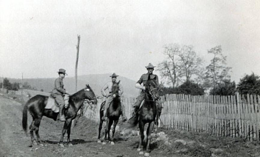 Three cadets atop three horses riding on a road parallel to a fence