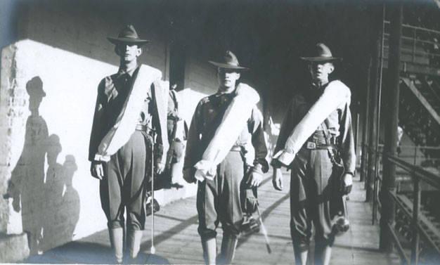 Three cadets standing within barracks wearing a long white sash, pants tucked into their socks, and wide brimmed hats