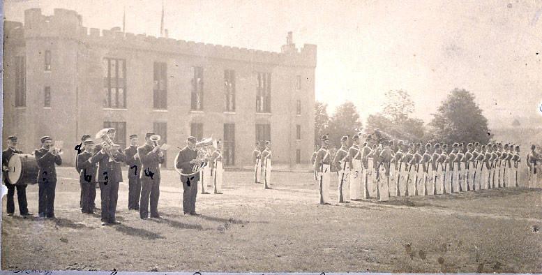 In the background barracks, in the foreground on the left a group of uniformed personnel playing tubas and a drum set. On the right a formation of cadets arranged in their coatees
