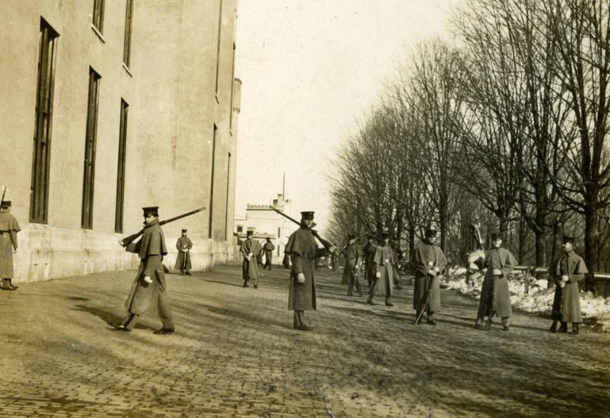 A group of cadets arranged outside of barracks carrying rifles. They walk in between barracks on the far left and a row of trees on the right.