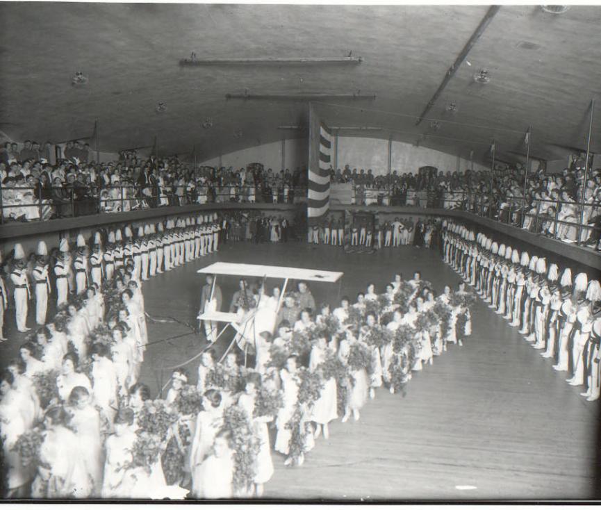 Cadets arranged around the outside of a square gymnasium with an American flag at its center. In the center of the floor stands an arrow formation of young girls wearing dresses and holding flowers.