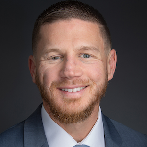 promotional portrait of Kyle Carpenter, white male with reddish hair, moustache, and trimmed beard in grey suit with white shirt and blue tie