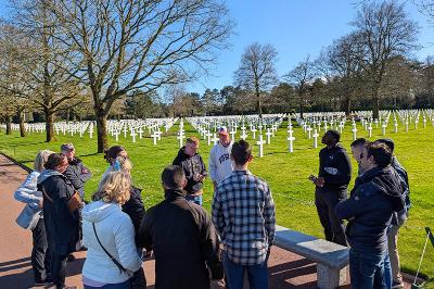 Cadets and VMI faculty leaders stand and talk at the Normandy American Cemetery, with the distinct white cross markers in the background.
