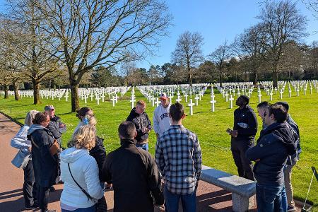 Cadets and VMI faculty leaders stand and talk at the Normandy American Cemetery, with the distinct white cross markers in the background.